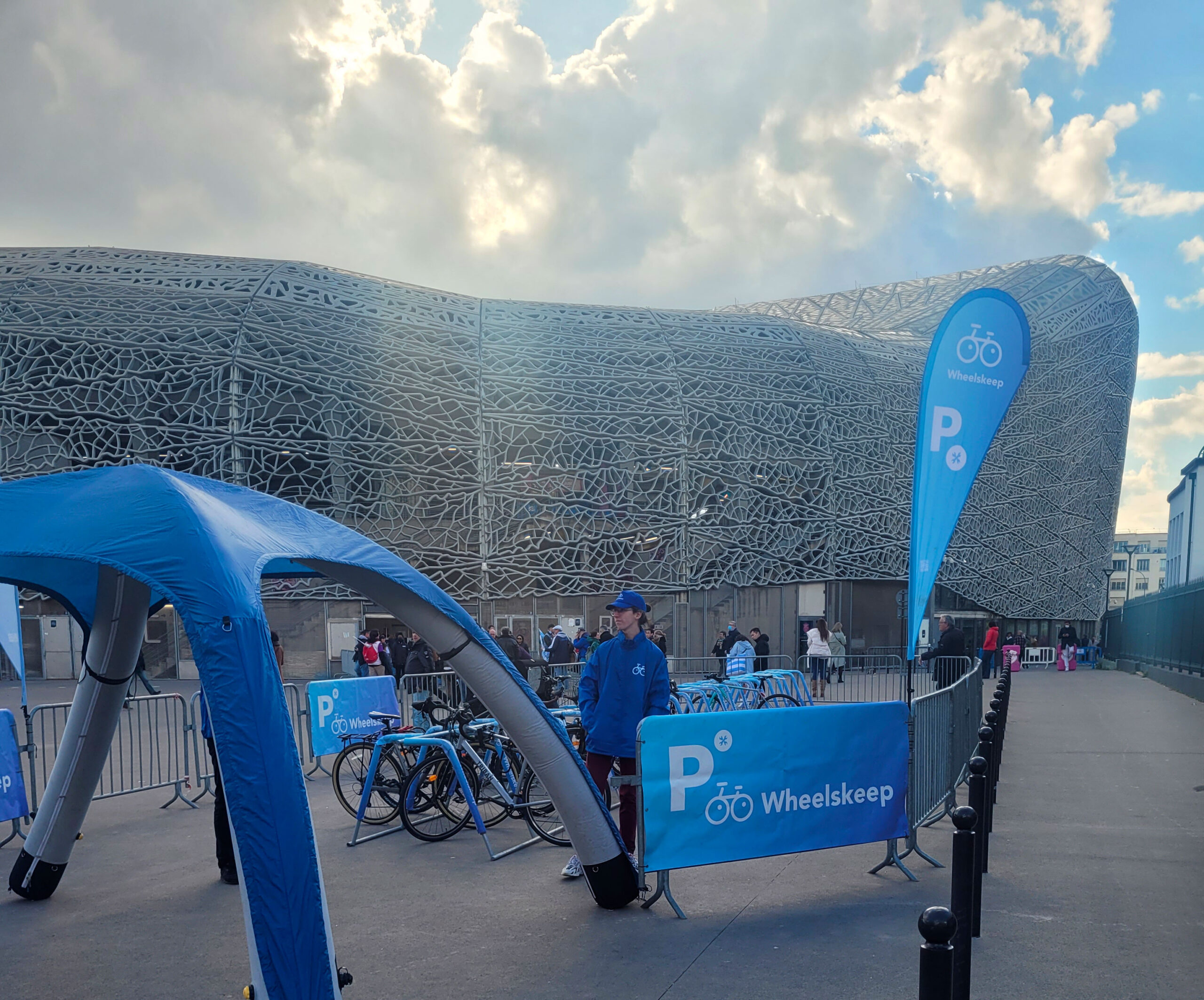 Bicycle rental area at an event, blue Wheelskeep banners, bikes lined up behind barricades near a lattice-pattern building.