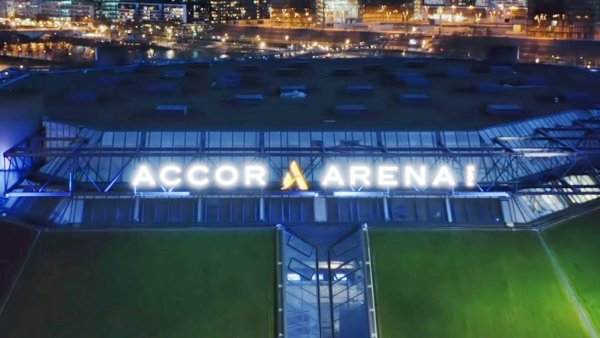 Aerial night view of the Accor Arena exterior with its illuminated sign and city lights in the background.