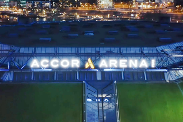 Aerial night view of the Accor Arena exterior with its illuminated sign and city lights in the background.