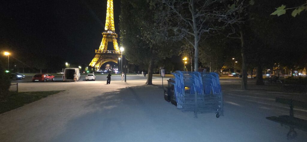 Night view of the Eiffel Tower lit in yellow, seen from a park with stacked blue chairs in the foreground.